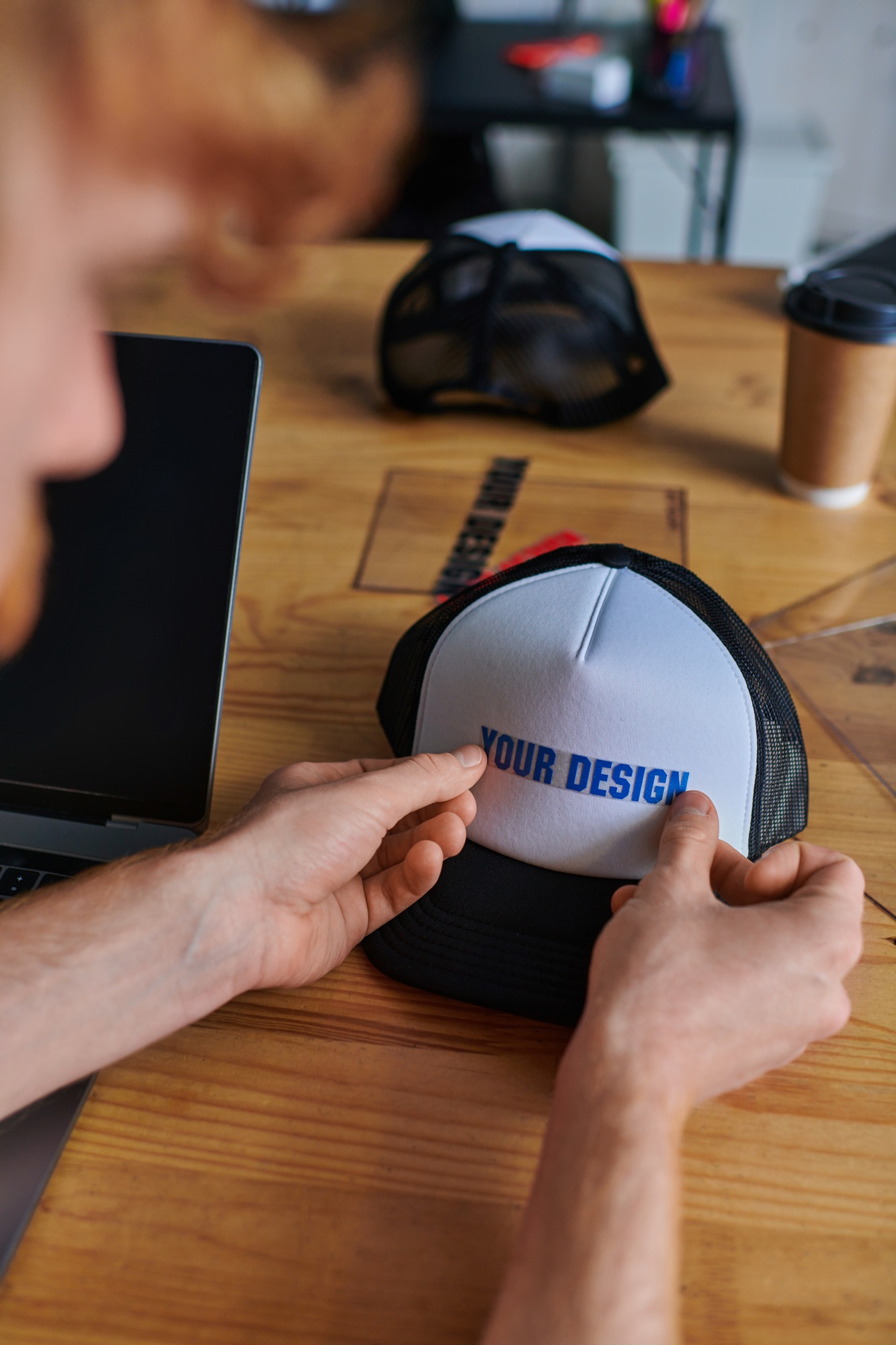 Cropped view of blurred young craftsman holding printing layer near snapback on table and laptop